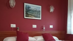 A neatly made bed with a floral bedspread, two red pillows, and a painting of a landscape on the wall. - Hotel Campus (Burgos)
