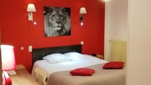 Habitación de hotel con pared roja, cama con ropa de cama blanca, dos almohadas rojas, un escritorio de madera con una lámpara y dos lámparas de pared.