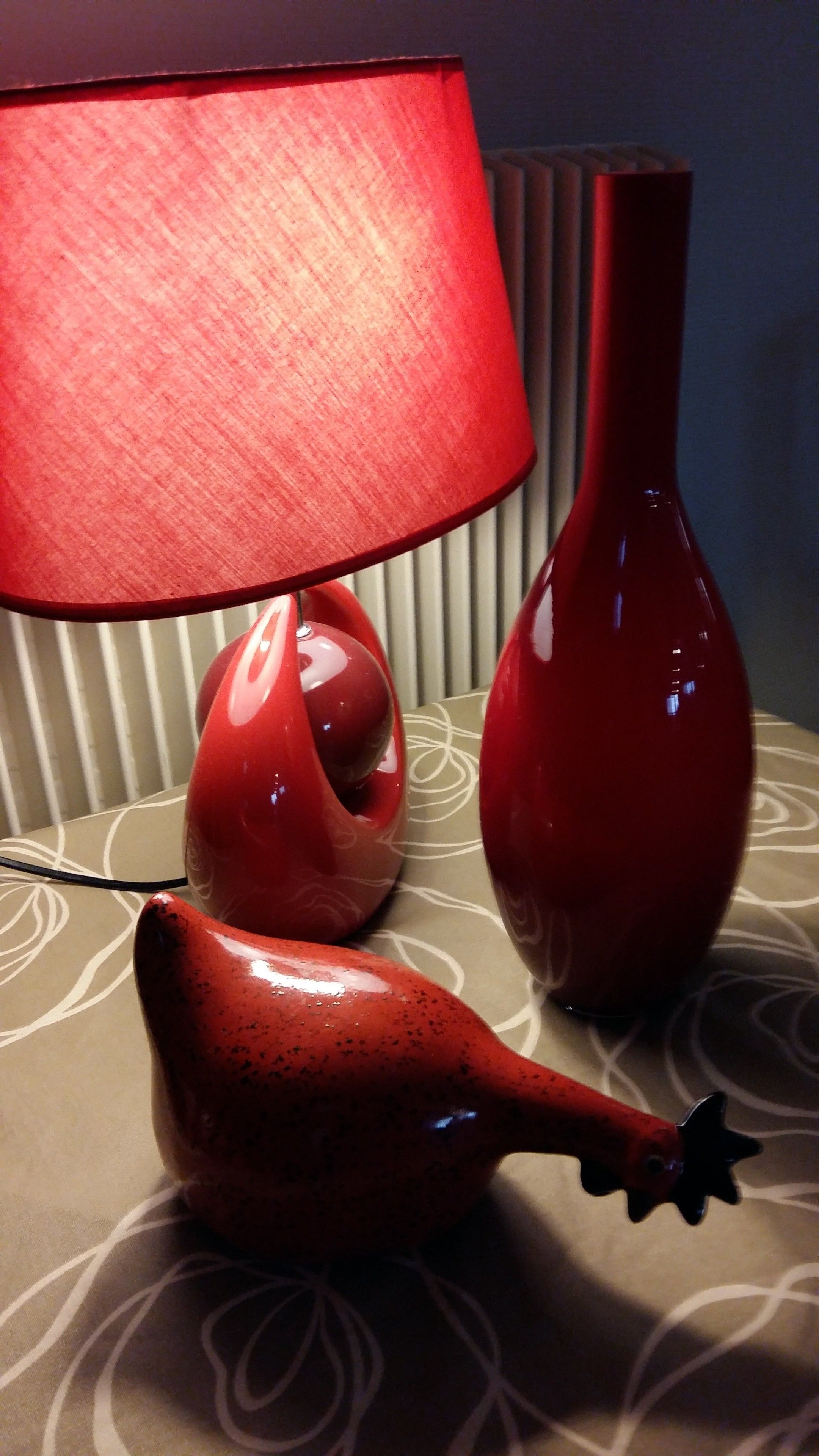 A lamp with a red shade, a red vase, and a red sculpture on a patterned table.