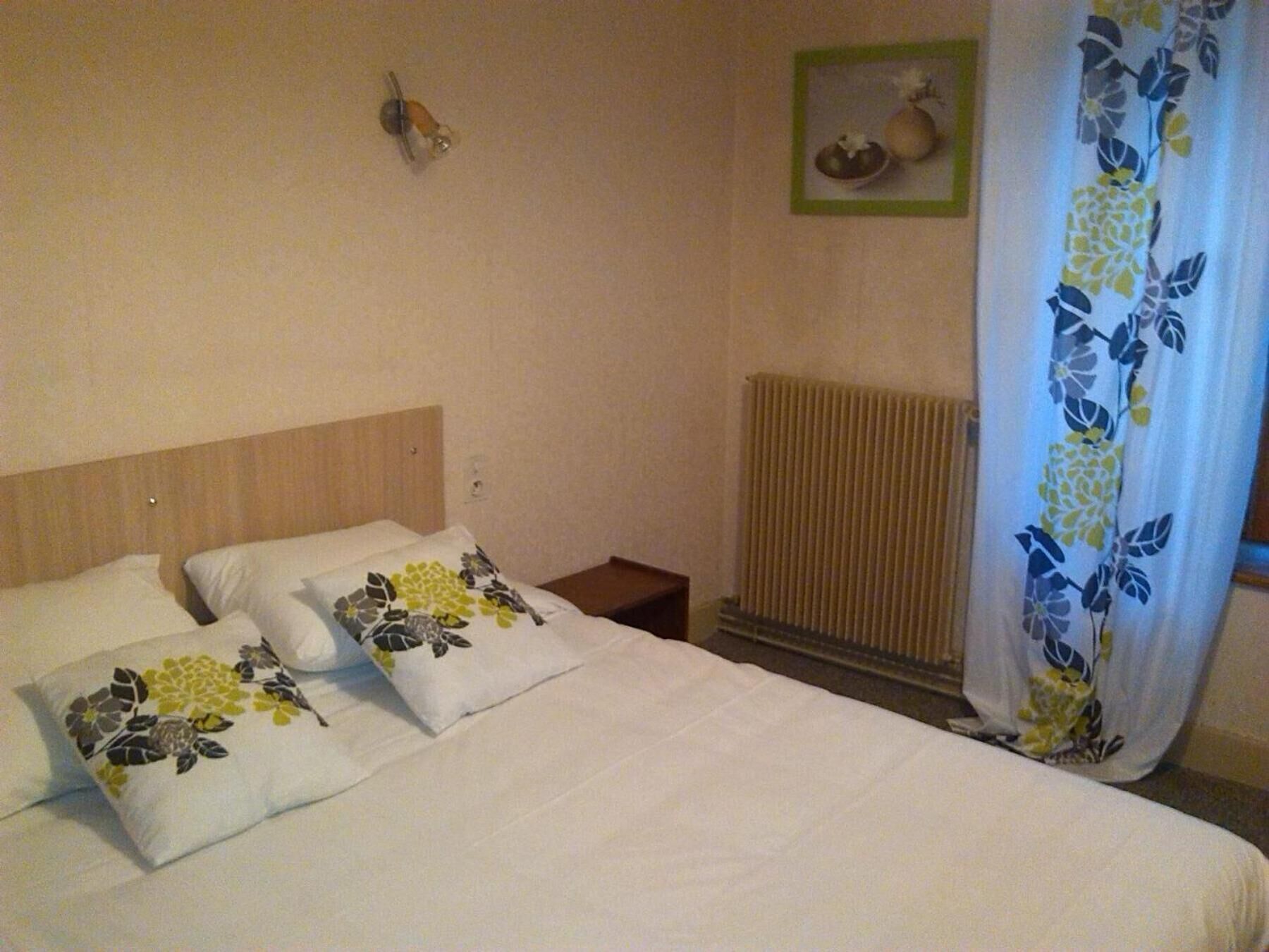 Habitación de hotel con cama, dos almohadas con motivos florales, mesita de noche, lámpara de pared, cuadro enmarco, radiador y cortina estampada con flores.