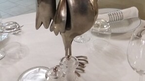 Una mesa de comedor con un centro de mesa decorativo de un gallo plateado, rodeada de copas y platos de cristal.