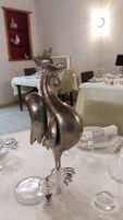 Uma mesa de jantar com um centro de mesa decorativo de galo prateado, rodeada por copos e pratos de vidro transparente.