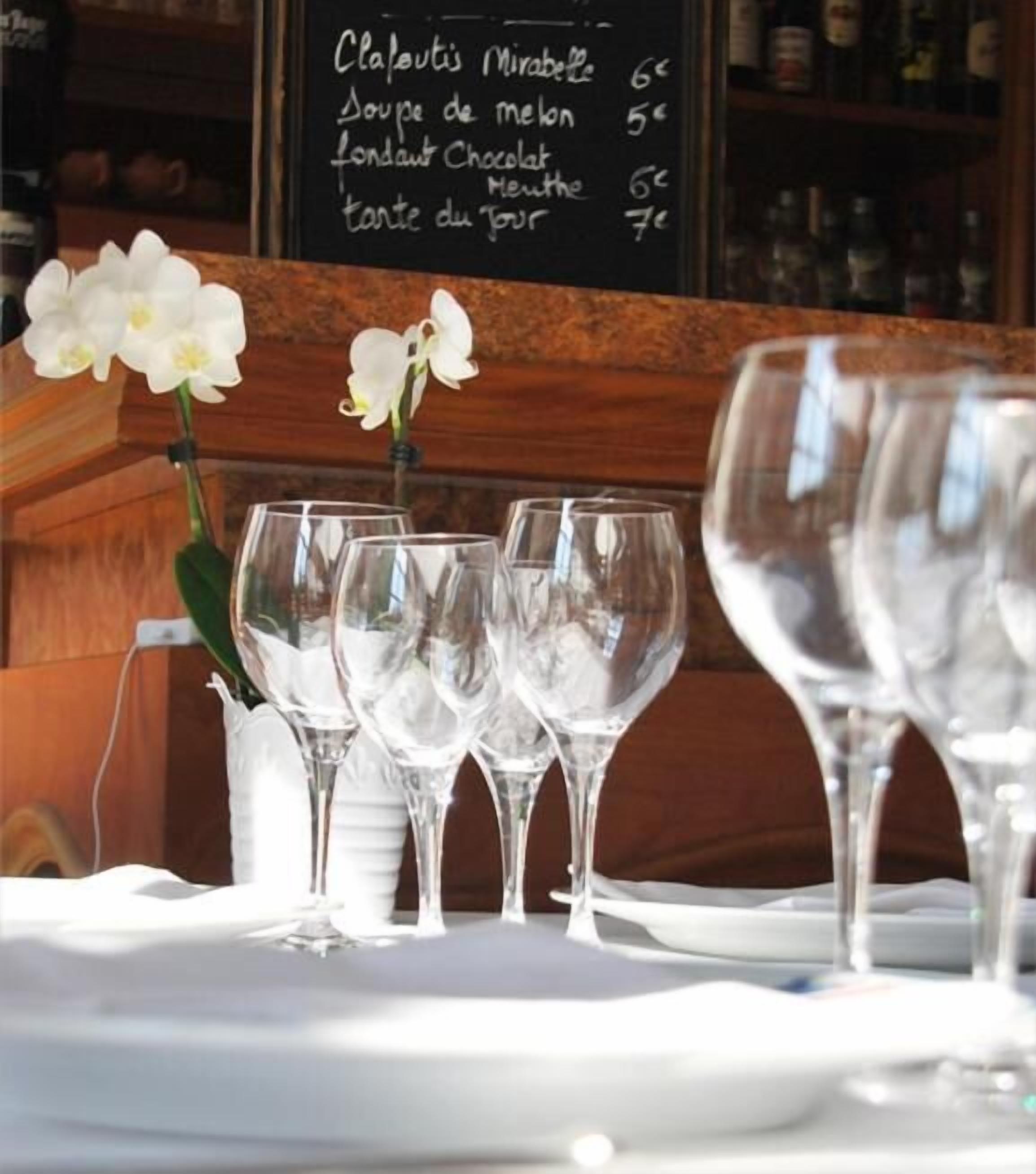 Une scène de restaurant avec, en arrière-plan, un tableau affichant le menu, où l’on peut voir des plats comme la clafoutis aux mirabelles et la soupe de melon.