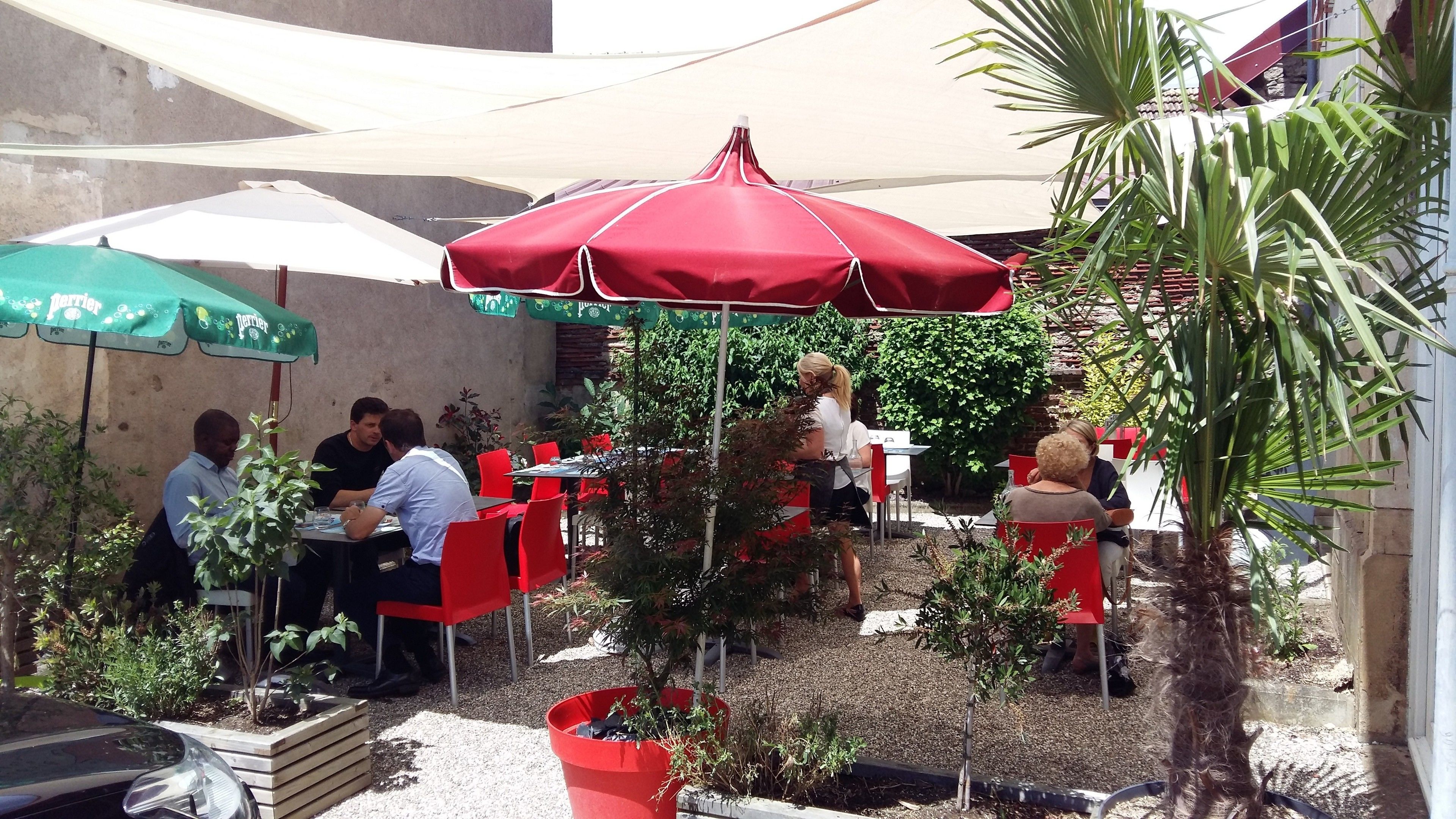 An outdoor seating area with tables and chairs under red and green umbrellas, surrounded by potted plants and a gravel ground.