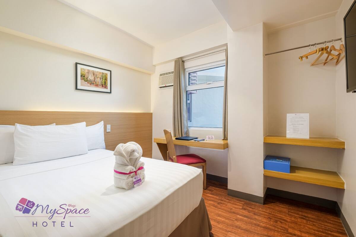 Habitación de hotel con cama, escritorio, silla, ventana y una estantería con una caja azul.