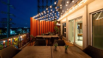 Un área de comedor al aire libre con mesas y sillas, luces colgantes y vista del paisaje urbano al anochecer.