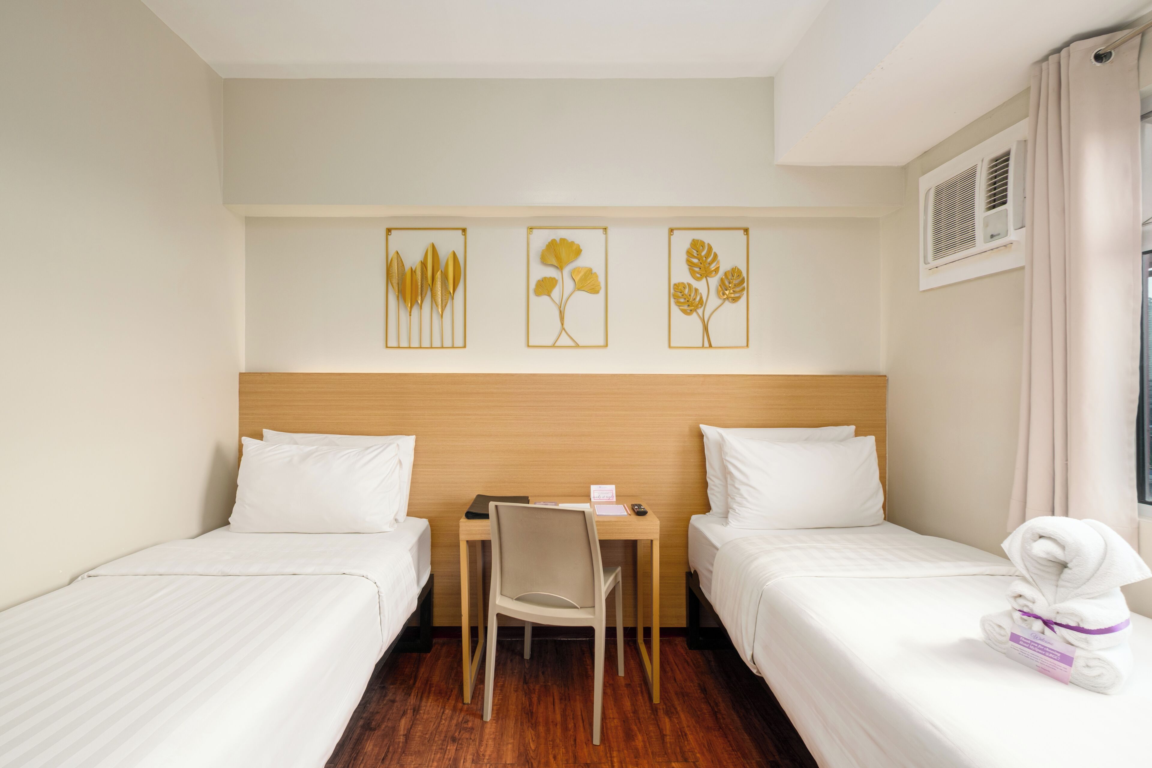 A hotel room with two single beds, a wooden headboard, a desk with a chair, and three wall-mounted artwork pieces.