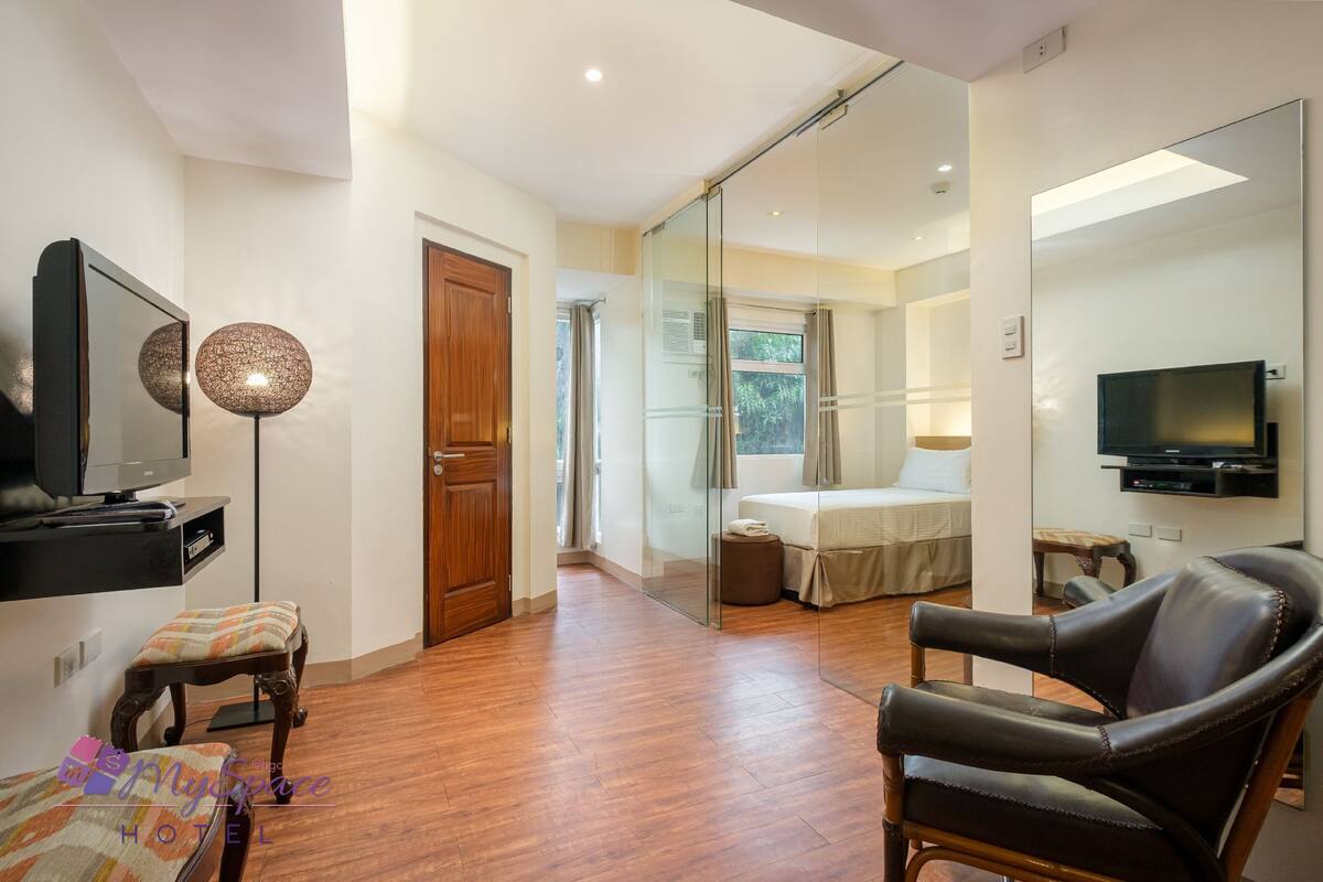 Una habitación de hotel moderna con suelo de madera, televisión, sofá, sillón, cama y un espejo grande.
