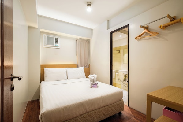Une chambre d’hôtel avec un lit, une table de chevet en bois, un porte-serviettes mural, une fenêtre avec des stores et une salle de bain visible à travers une porte ouverte.