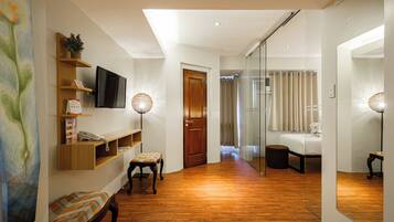 A hotel room with a wooden floor, a desk with a telephone, a chair, a bed, a mirror, and a wall-mounted TV.