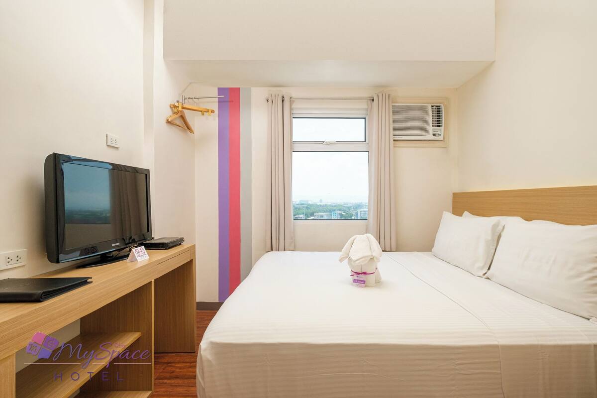 Habitación de hotel con una cama, televisión, escritorio y ventana con cortinas.