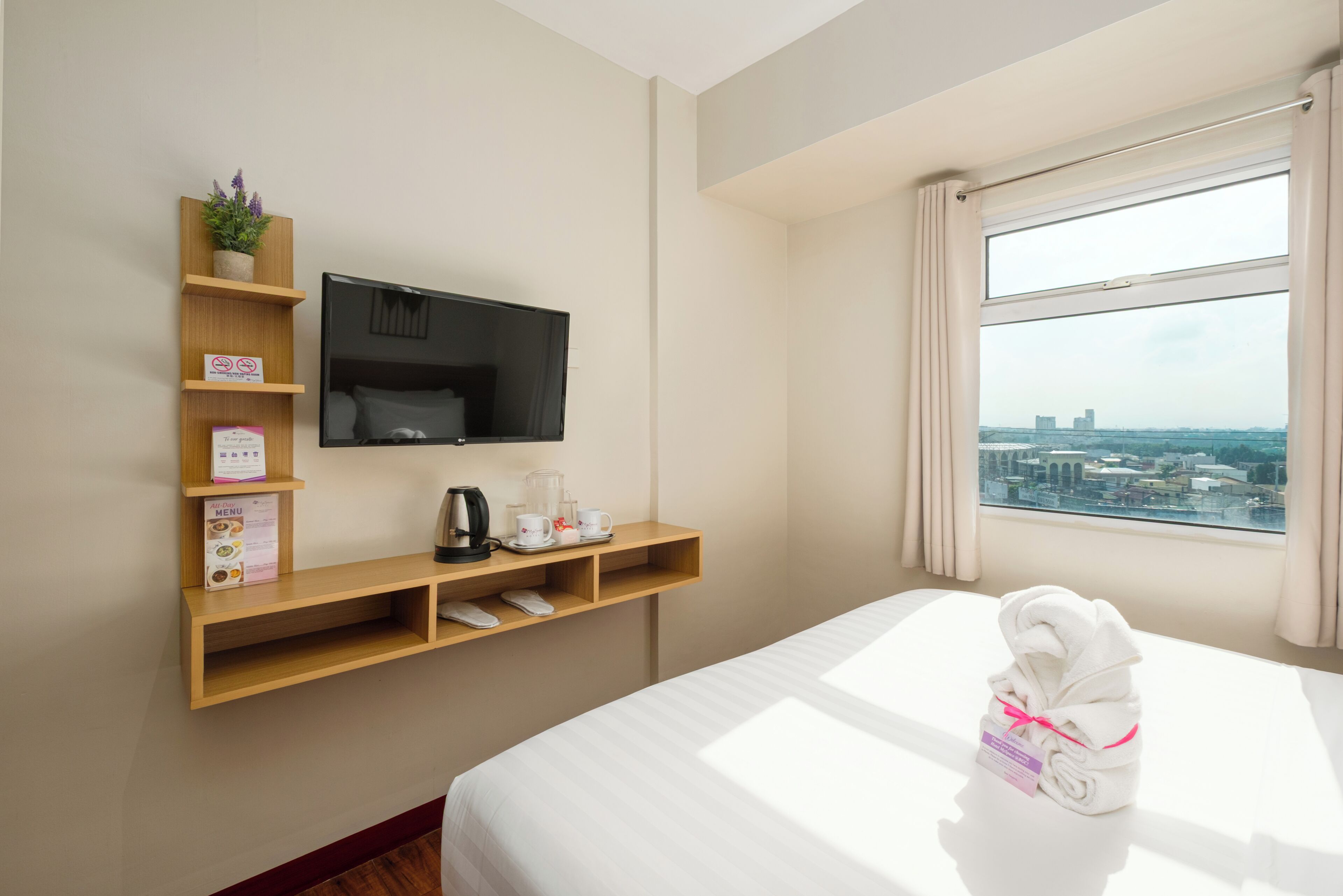 A hotel room with a bed, a TV mounted on the wall, a window with a city view, and a shelf with amenities.