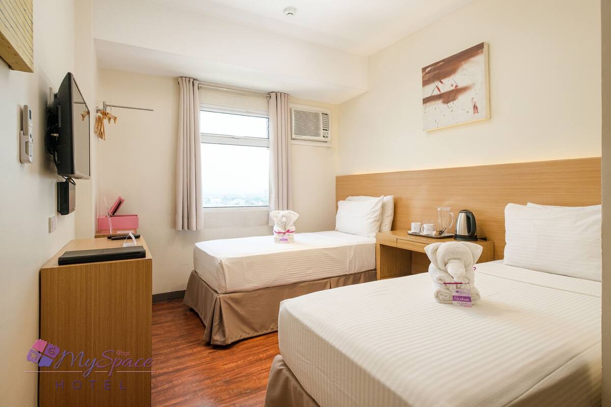 Habitación de hotel con dos camas, un escritorio con computadora, un televisor y una ventana con cortinas.