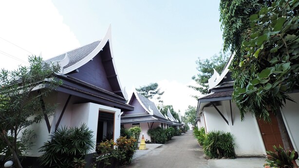 A row of traditional Thai-style buildings with steeply pitched roofs, surrounded by lush greenery and a paved pathway. - Sweet Inn Resort Hotel (Bang Pahan)