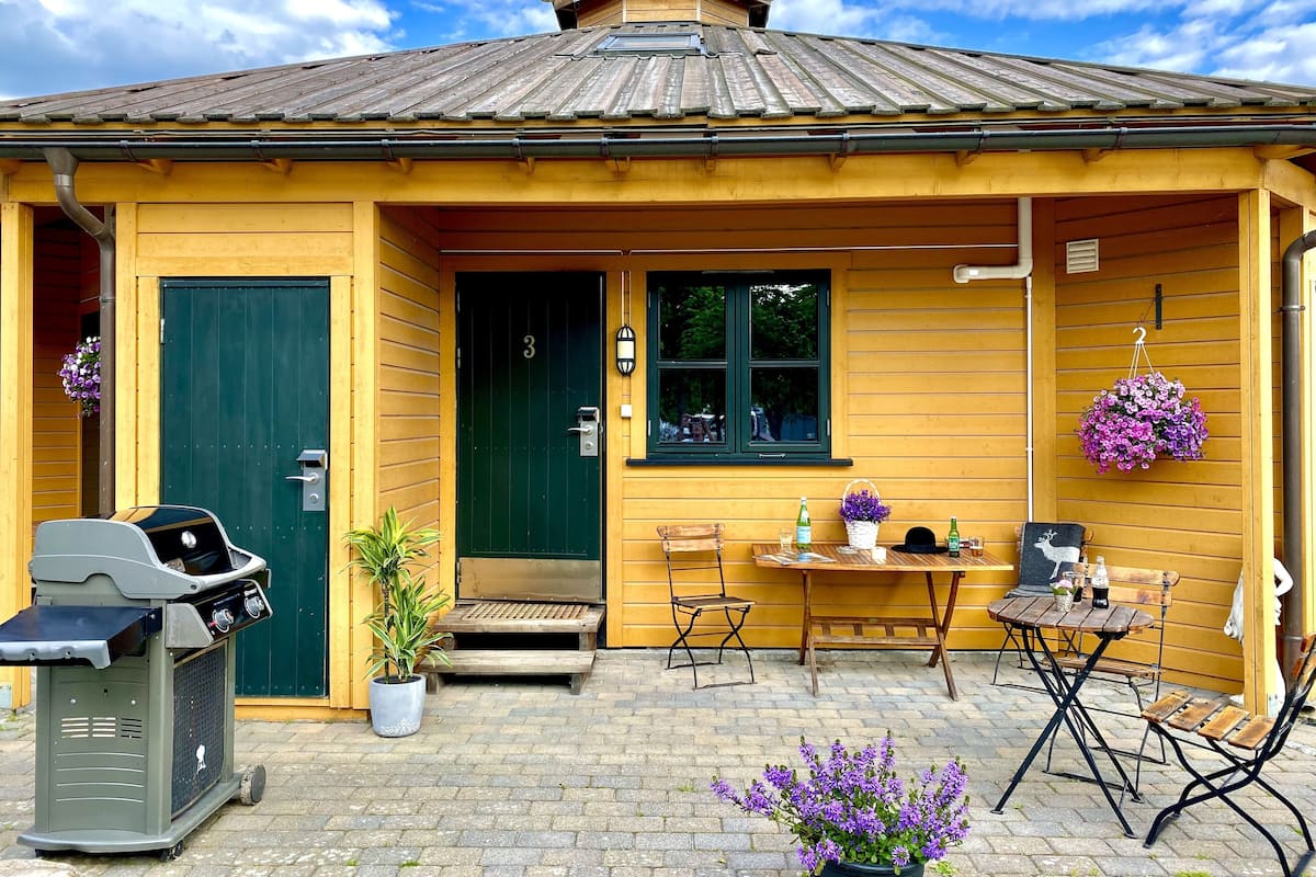 Una construcción de madera con una puerta verde, un pequeño porche con una mesa y sillas, y una parrilla de barbacoa al exterior.