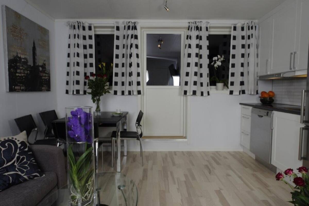 Una cocina moderna con armarios blancos, una zona de comedor con mesa de vidrio y una sala de estar con sofá y un jarrón decorativo.