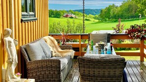 Une terrasse en bois, meublée de meubles en osier, ornée d’une statue et offrant une vue sur un paysage verdoyant.