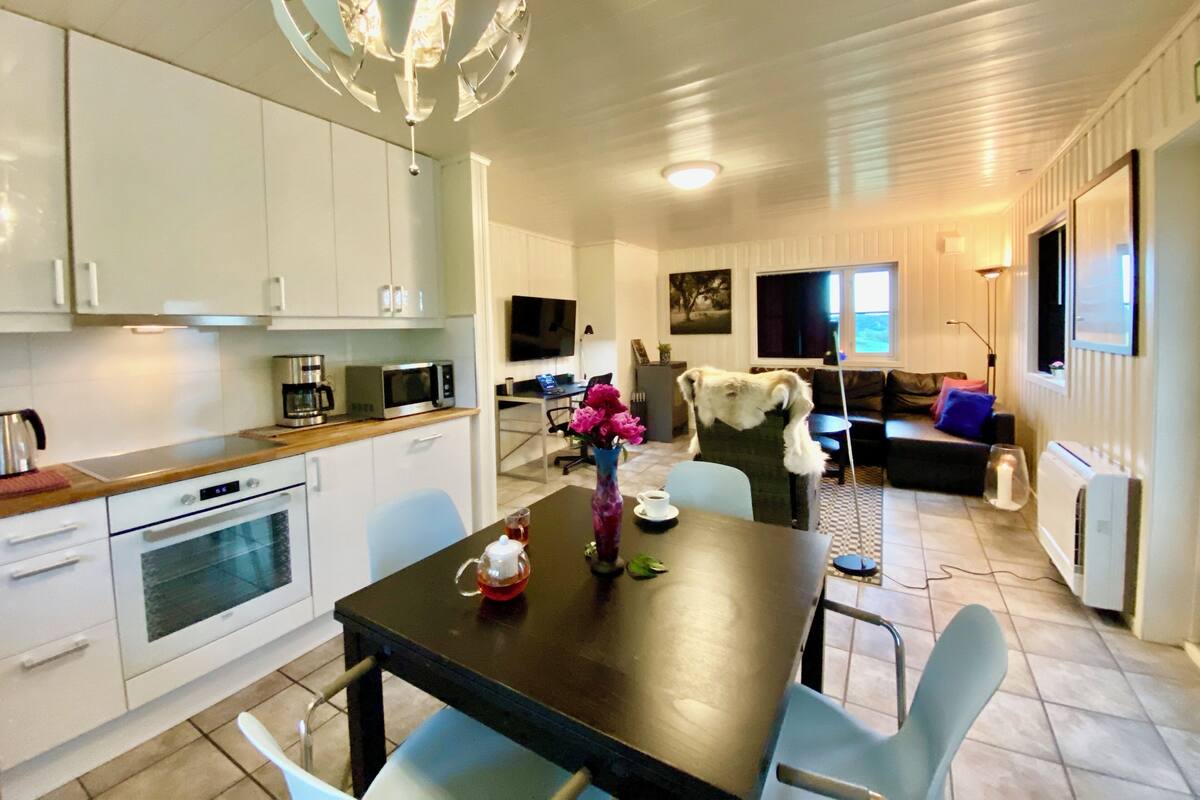 Una cocina moderna con armarios blancos, microondas y cafetera. Hay una mesa de comedor con sillas y un jarrón de flores.