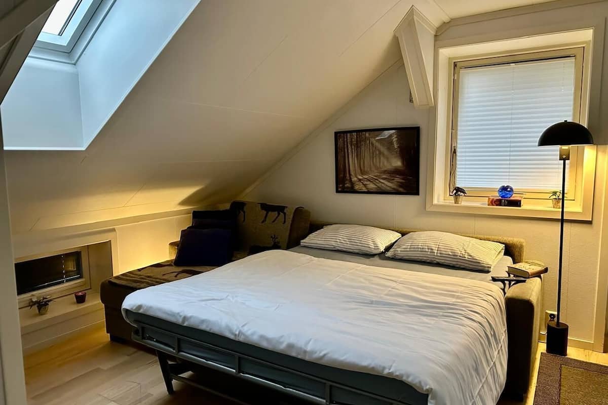 Un dormitorio con techo inclinado, una cama, una mesita de noche, una lámpara, una ventana con persianas y una repisa con libros y un globo terráqueo.