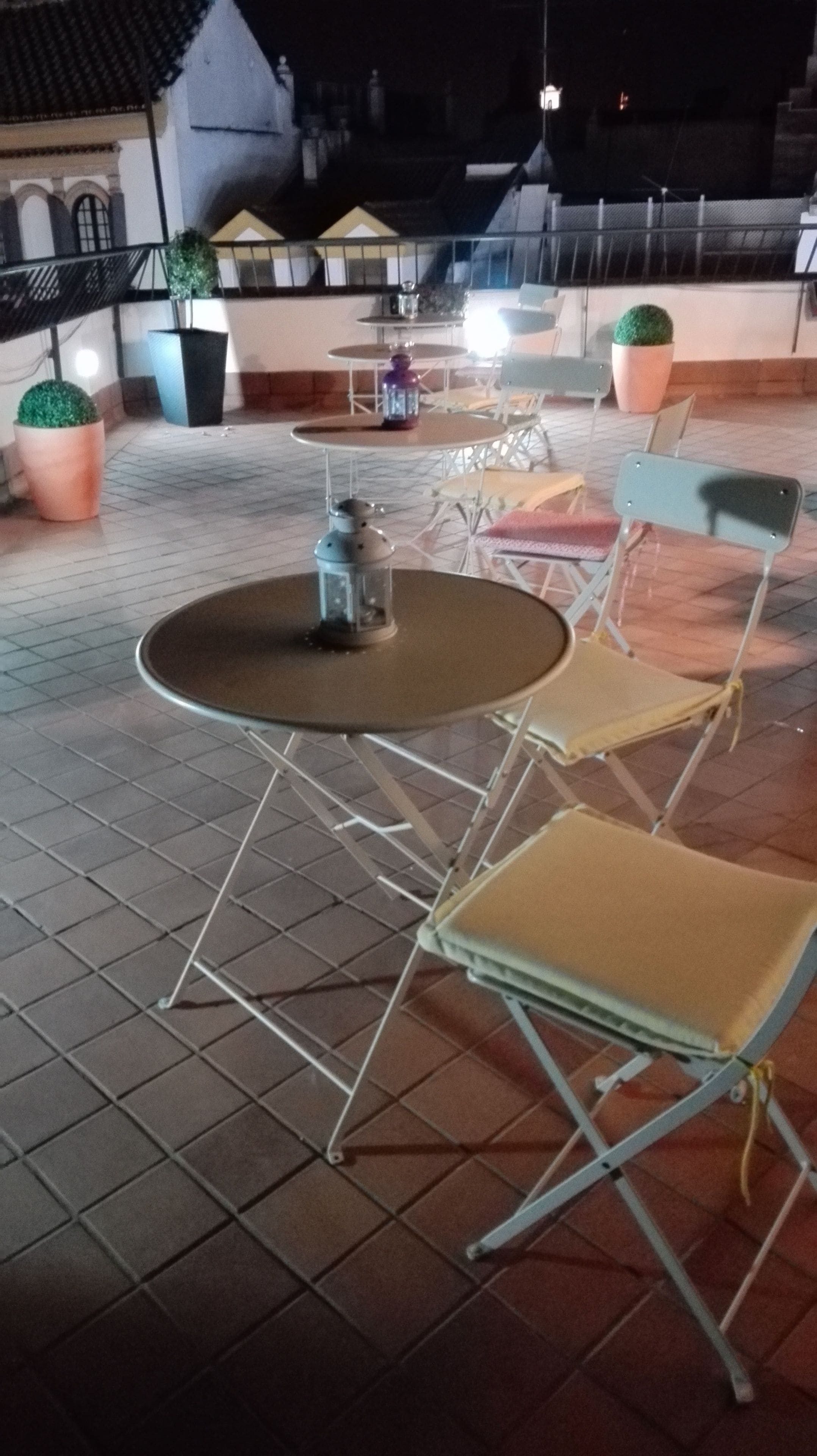 Un espacio al aire libre con mesas, sillas, plantas en macetas y piso de baldosas.