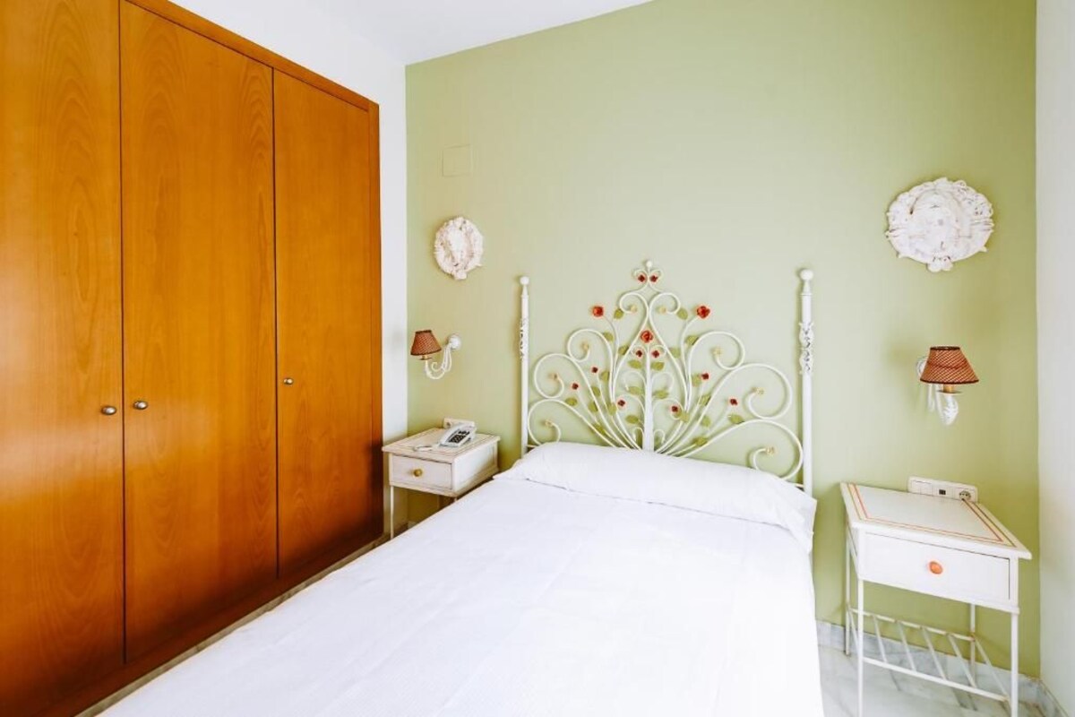 A bedroom with a wooden wardrobe, a bed with a white headboard, bedside tables with lamps, and decorative wall pieces.