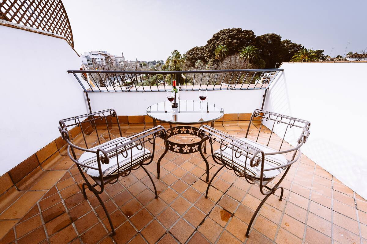 A terrace with a round table and two metal chairs, a bottle of wine, and a view of buildings and trees.