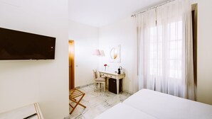 A hotel room with a bed, a desk with a chair, a TV mounted on the wall, a mirror, and a window with curtains. - Basic Hotel Doña Manuela (Seville)