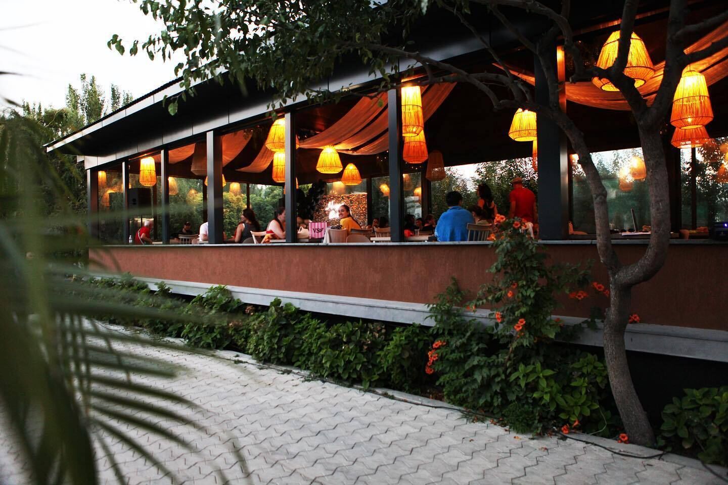 Un restaurante al aire libre con zona de estar techada, ventanas amplias y faroles colgantes.