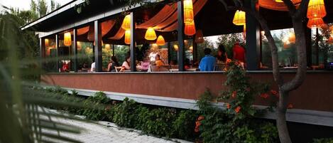 Un restaurante al aire libre con zona de estar techada, ventanas amplias y faroles colgantes.