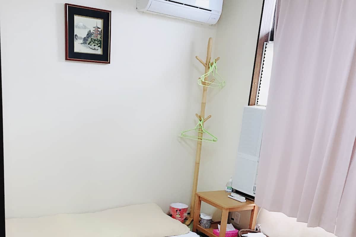 Une chambre avec un lit, une petite table, un climatiseur, un tableau encadré et un porte-vêtements suspendu.