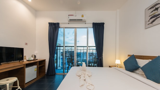 A hotel room with a bed, a desk with a television, a mini-fridge, a balcony with a view of the city, and a ceiling air conditioner. - Two Color Patong (Patong)