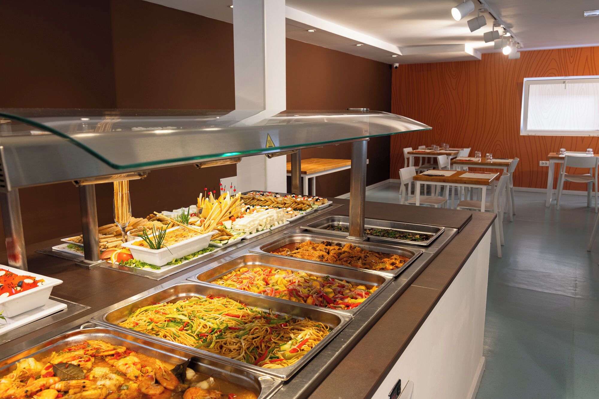 A buffet with various food options, including seafood, noodles, and salads, in a modern dining area with tables and chairs.