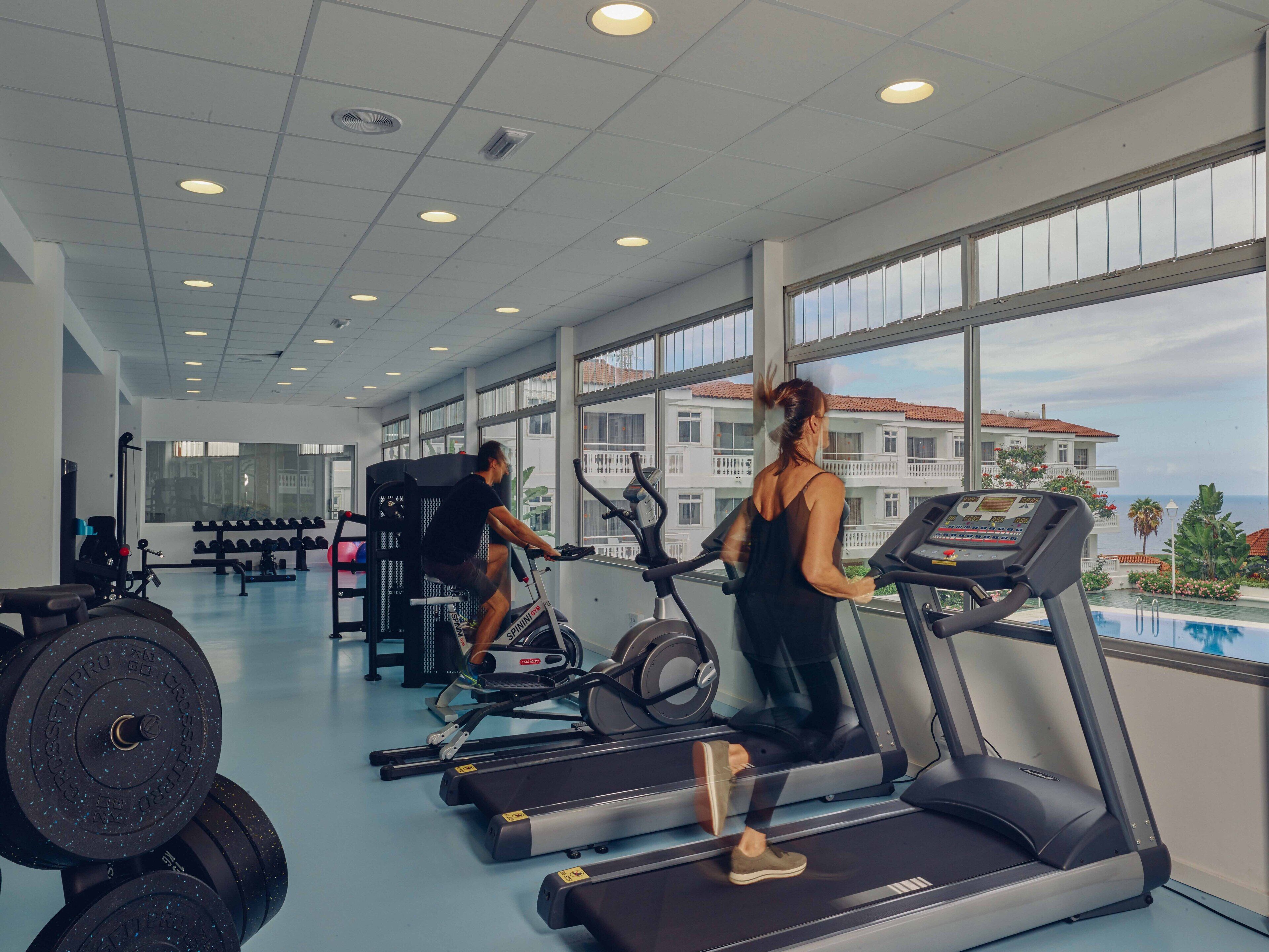 Un gimnasio con cintas de correr y equipo de pesas, ventanas amplias y vistas a un edificio con balcones.