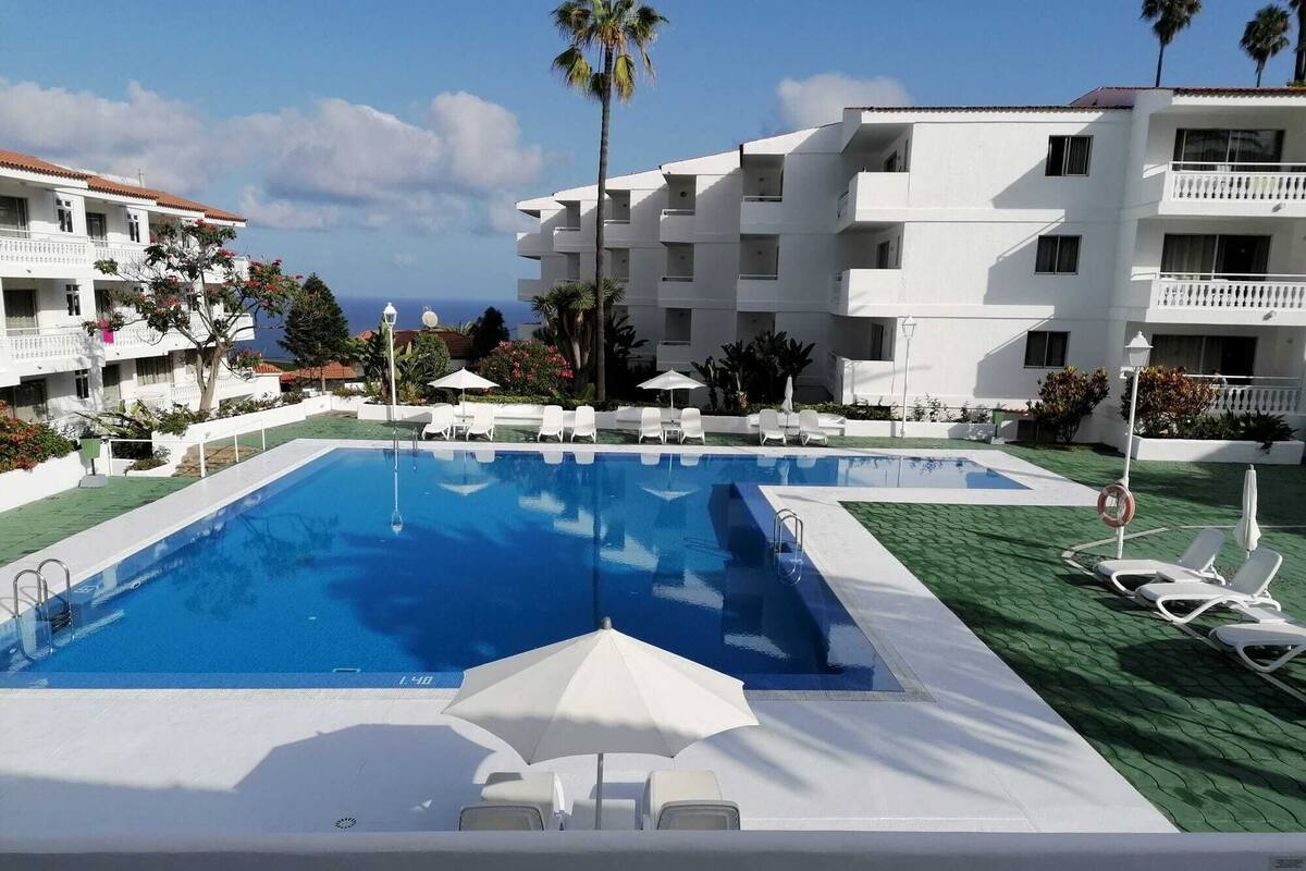 A swimming pool with lounge chairs and umbrellas, surrounded by white buildings with balconies, under a clear sky.