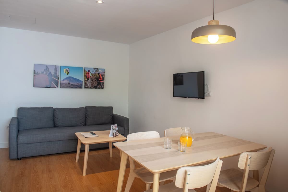A modern living room with a grey sofa, a wooden dining table with chairs, a TV mounted on the wall, and a pendant light.
