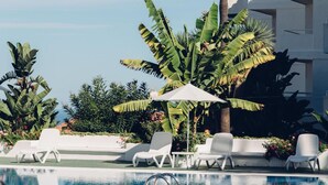 A pool area with lounge chairs and umbrellas, surrounded by palm trees and a building with balconies. - Route Active Hotel (Los Realejos)