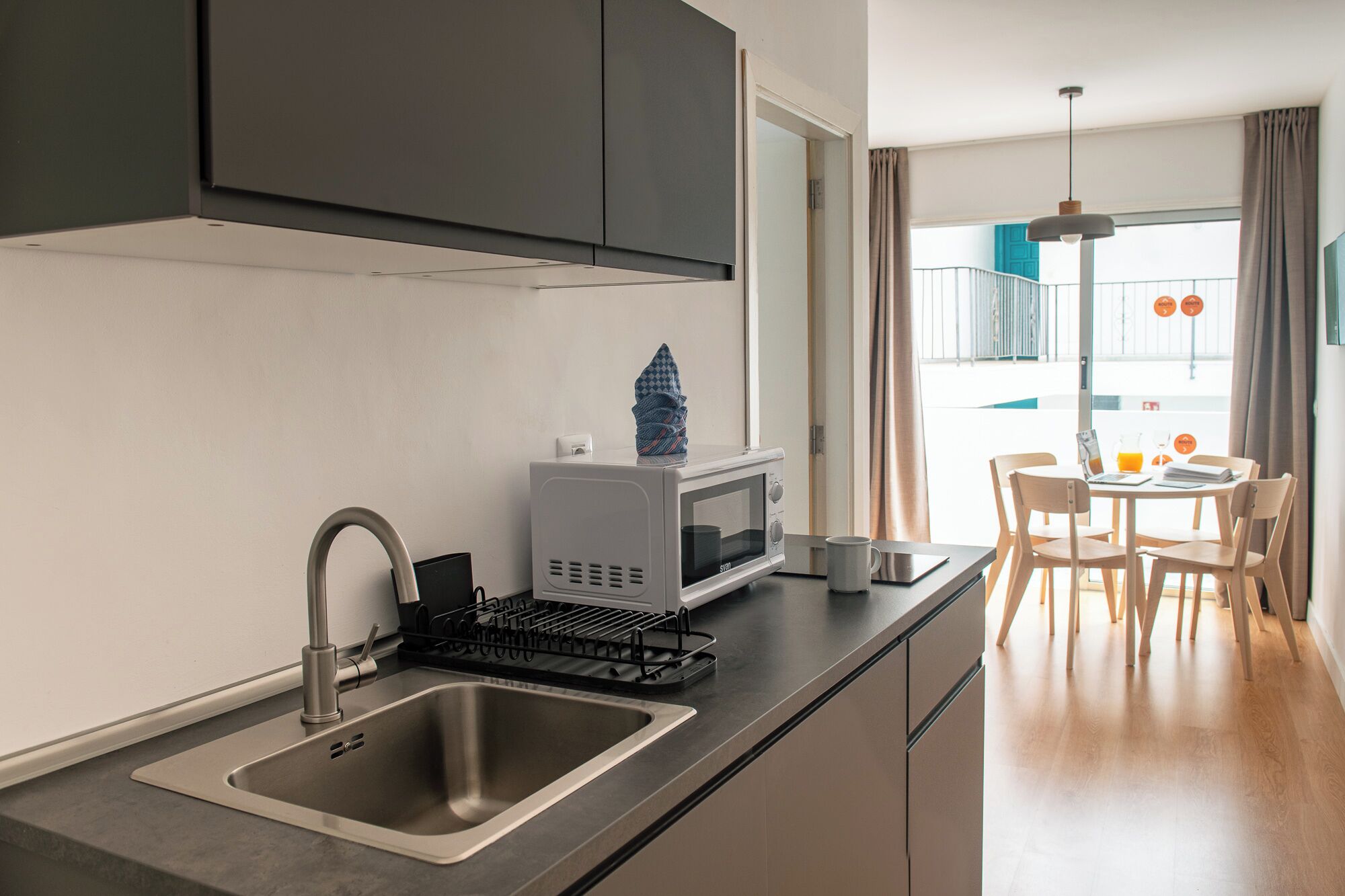 Una cocina moderna con pileta, microondas y tetera, un comedor con mesa y sillas, y vista al balcón exterior.