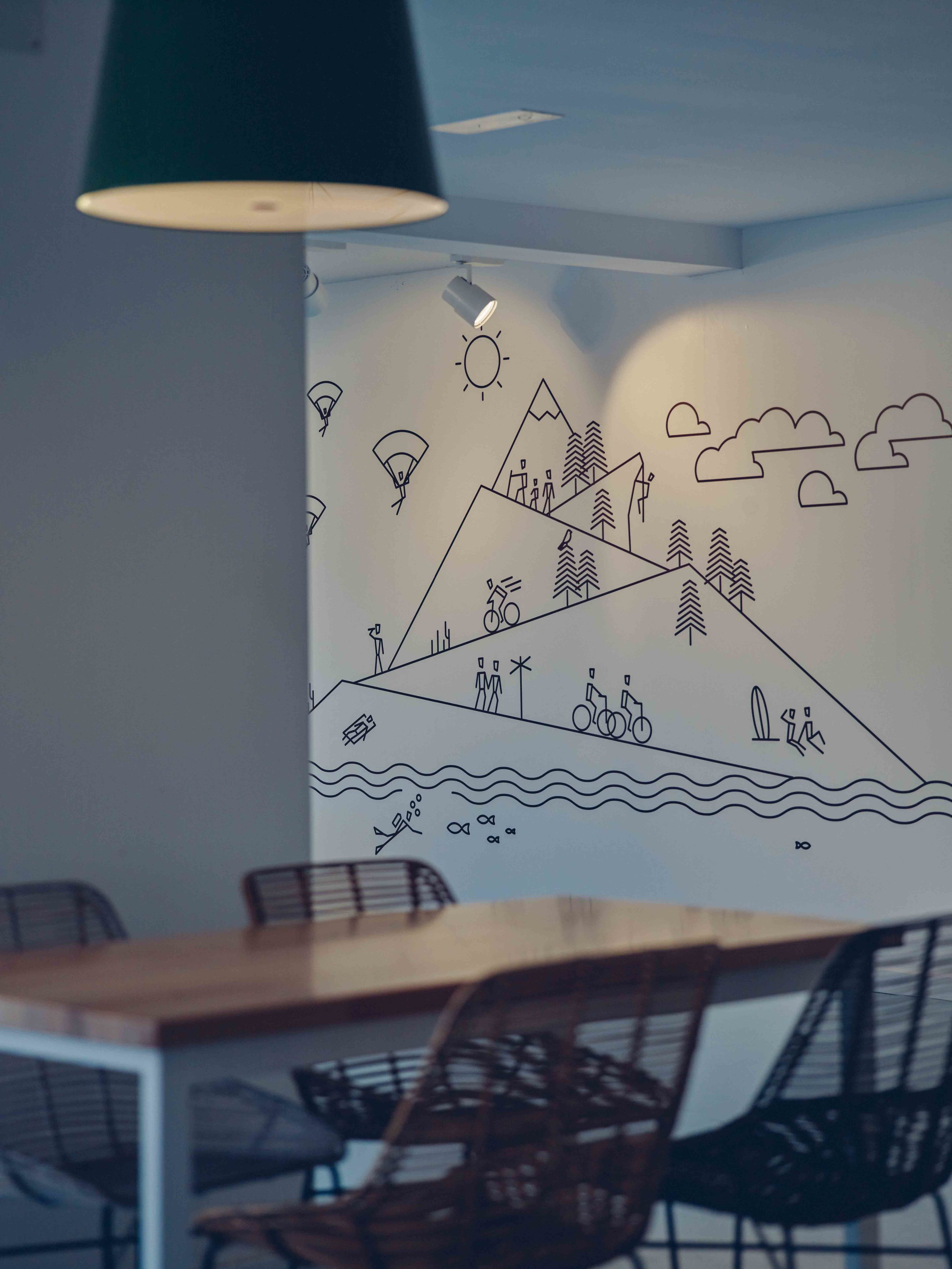 A dining area with a wooden table and wicker chairs, a wall mural depicting a mountain landscape with people and activities, and a hanging green pendant light.