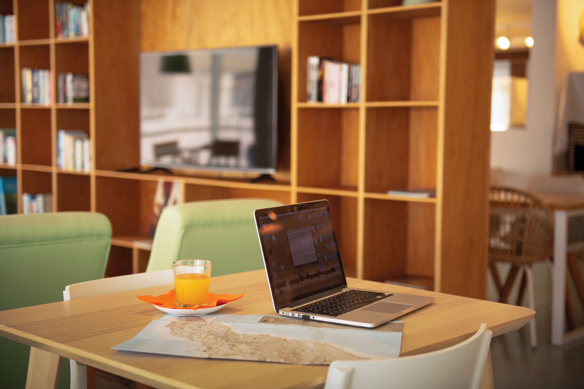 A laptop on a table with a glass of orange juice, a map, and a television in the background.