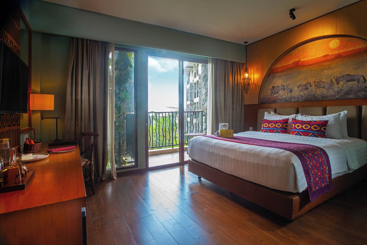 Habitación de hotel con una cama grande, un escritorio con lámpara, una silla y vistas al exterior a través de una puerta corredera.