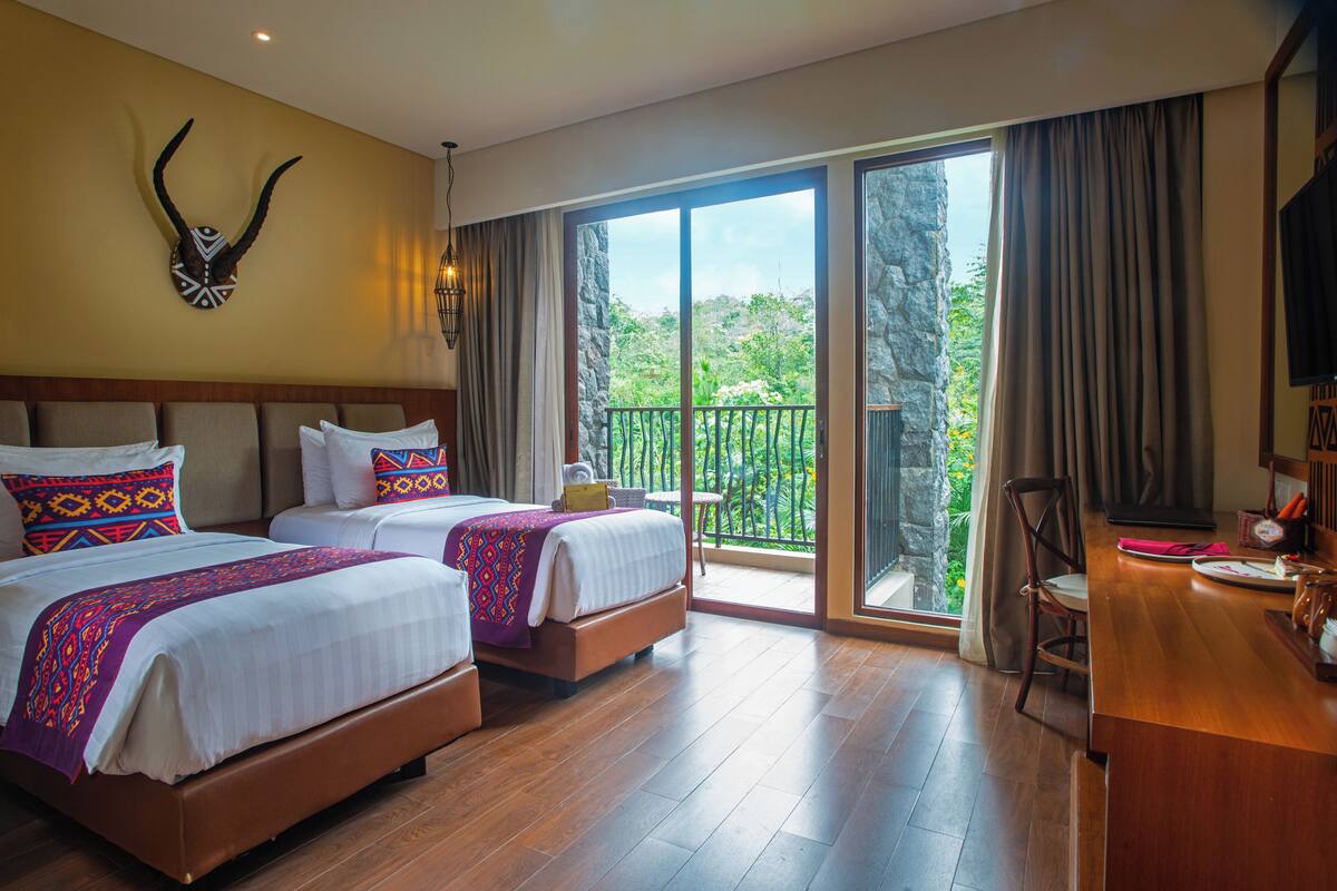 Habitación de hotel con dos camas, un balcón con vistas y un escritorio de madera con una silla.