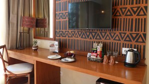 Habitación de hotel con escritorio de madera, televisor, sillón, planta, frutero, pastel y cafetera.