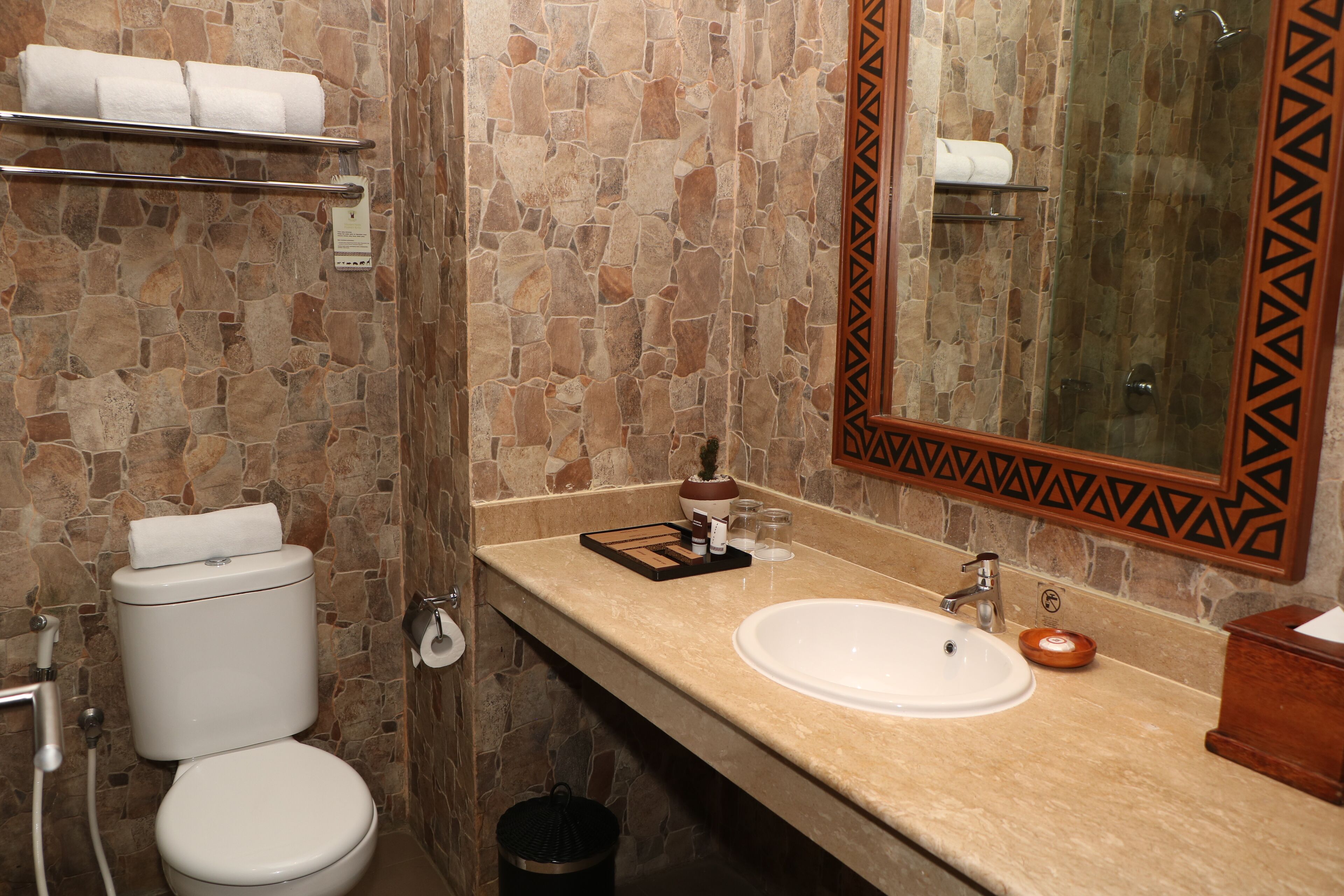 Ein Badezimmer mit Steinwand, Toilette, Waschbecken und einem Spiegel mit geometrischem Rahmen.