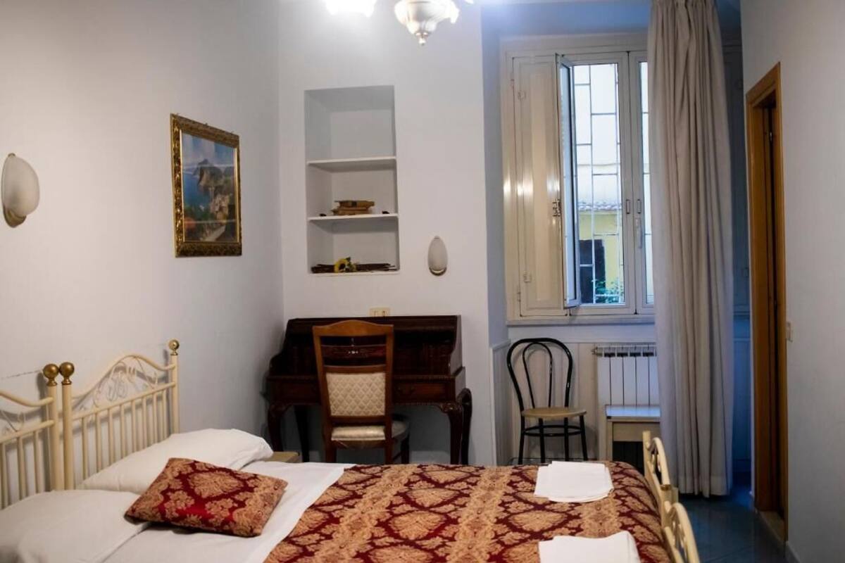 A bedroom with a bed, a desk, a chair, a radiator, and a window with curtains.
