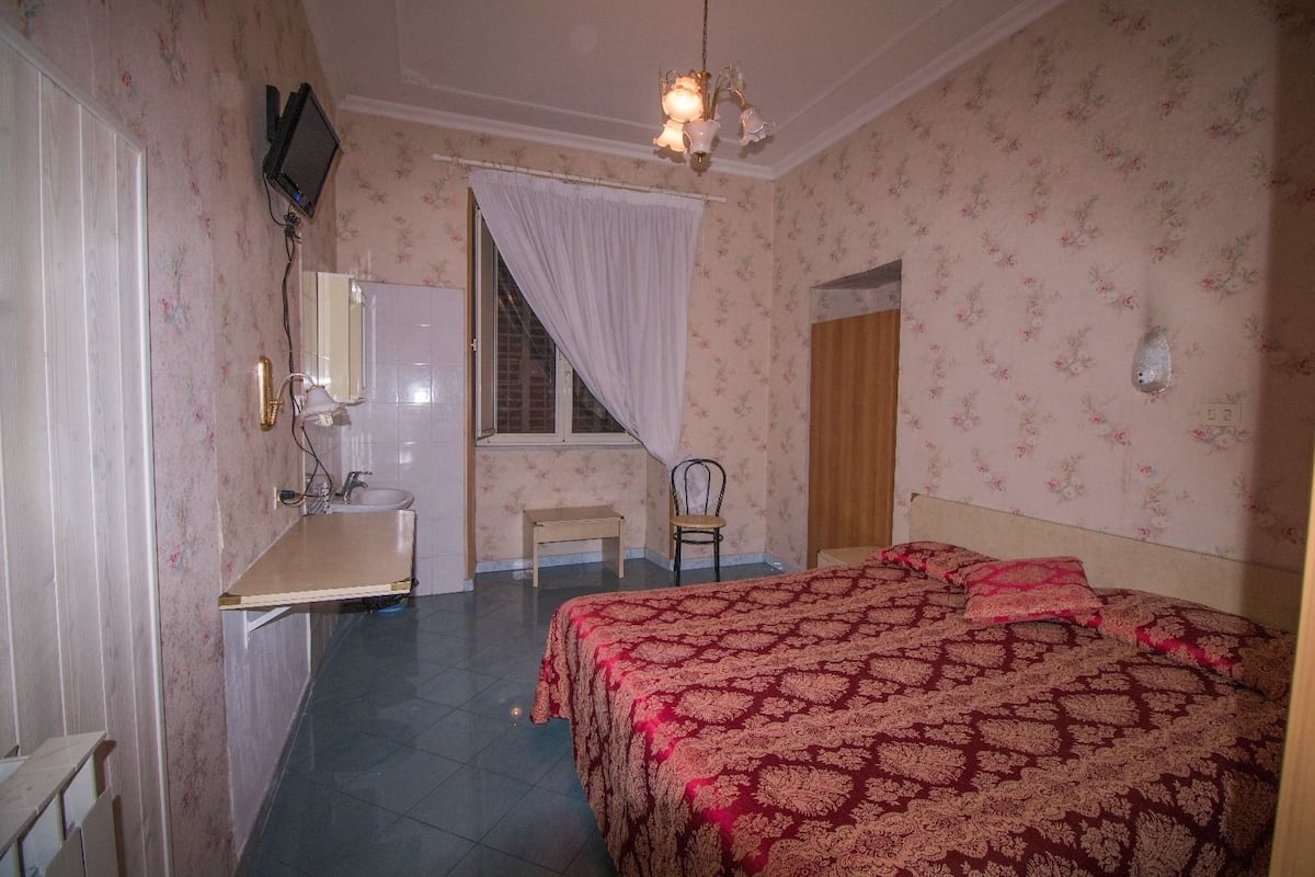 A bedroom with a bed, a chair, a desk with a mirror, a window with curtains, and a television mounted on the wall.