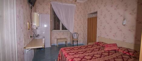 Un dormitorio con cama, una silla, un escritorio con espejo, una ventana con cortinas y un televisor fijado a la pared.