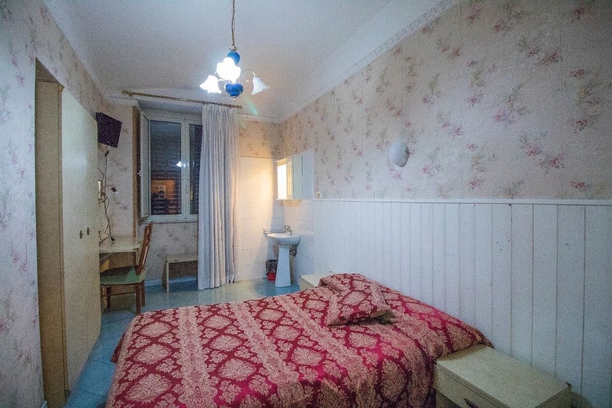 A small, dimly lit room with a bed, a chair, a small table, a window with curtains, and a wall-mounted mirror.