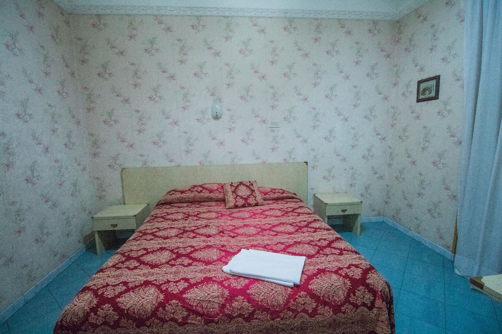 Makuuhuoneessa on kukkakuviollinen tapetti, punavalkoinen ja kultainen peitto sängyllä, kaksi yöpöytää ja pieni taulu seinällä.
