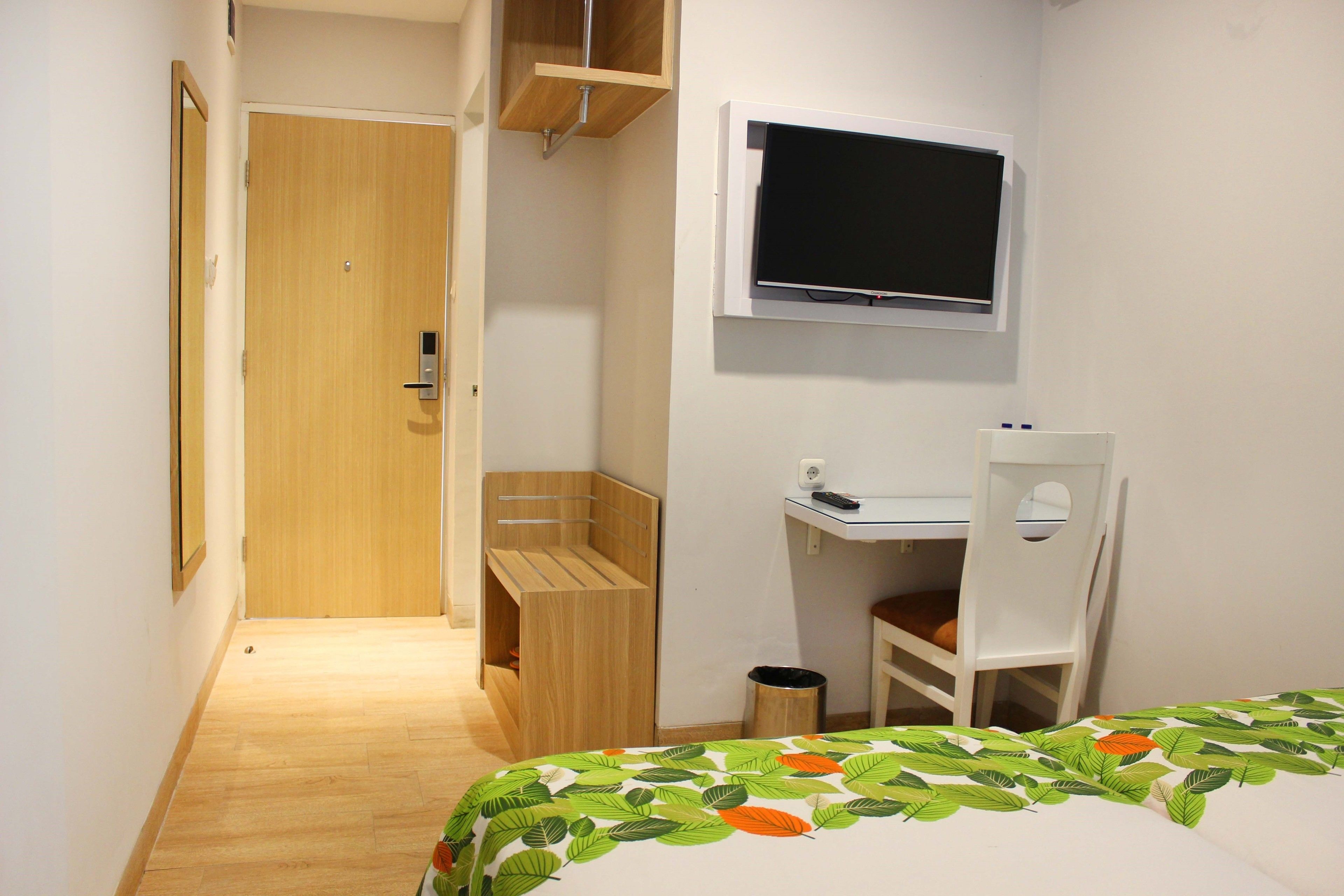 A small, well-lit room with a wooden door, a bed with a leaf-patterned bedspread, a white desk with a chair, and a mounted television.
