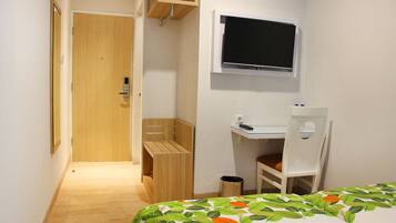 A small, well-lit room with a wooden door, a bed with a leaf-patterned bedspread, a white desk with a chair, and a mounted television.